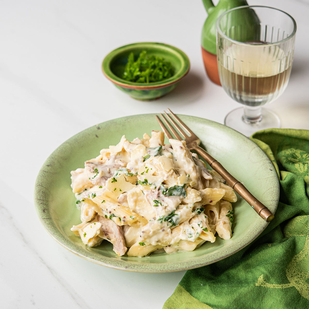 A plated pasta dish on a light green ceramic plate with a gold-toned fork, set on a white marble surface with a green napkin, a small green bowl of herbs, and a glass of white wine in the background.